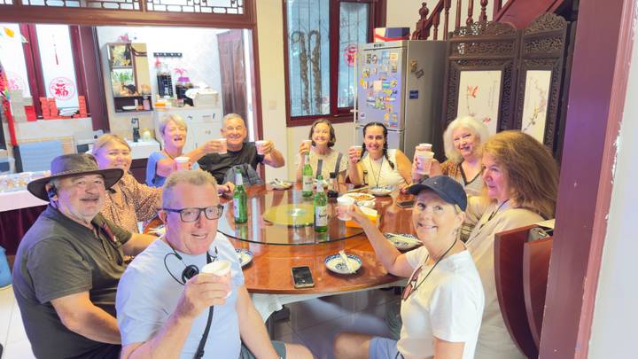 Group of travellers raise drinks around a circular dining table inside a traditional house.