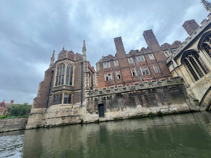 Historic college buildings rise beside the River Cam on an overcast day.