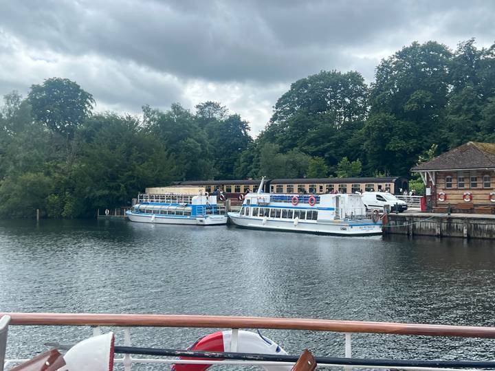 Docked sightseeing boats rest on calm lake waters bordered by leafy trees under grey skies.