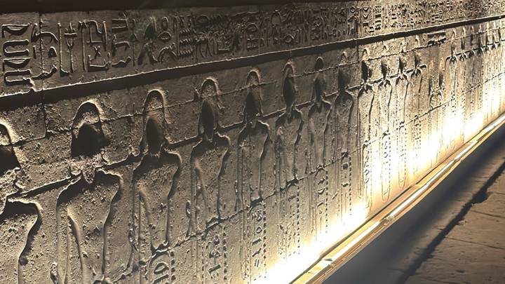 Dimly lit corridor of hieroglyphic wall reliefs illuminated by warm accent lighting.