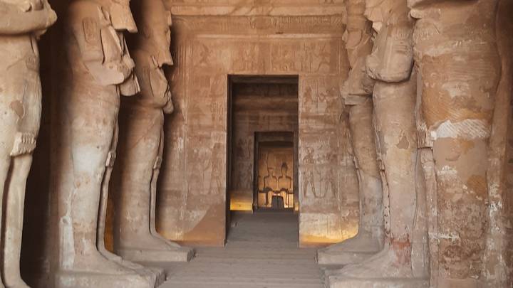 Interior corridor of an ancient Egyptian temple lined with towering sandstone statues and hieroglyphics.