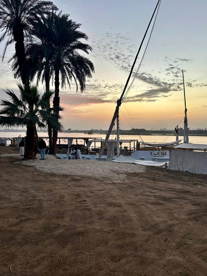 Traditional Nile sailboat moored at sandy riverbank at dawn, palms silhouetted against colorful sky.