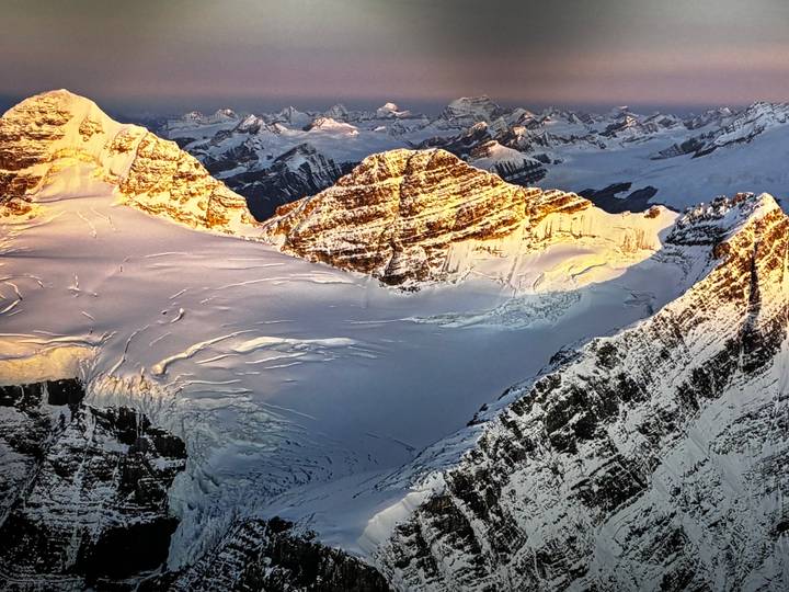 High-altitude view of snow-capped Rocky Mountain peaks glowing in sunrise light over a vast glacier.