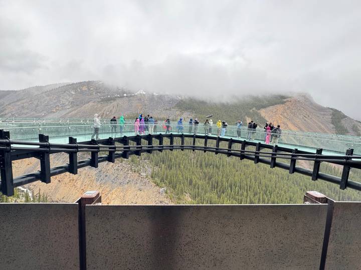 Glass skywalk spanning a canyon with visitors in colorful ponchos against misty mountain backdrop.