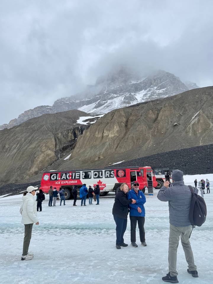 Large red Ice Explorer bus labeled 'Glacier Tours' parked on a snowy moraine with tourists milling about beneath towering cliffs.