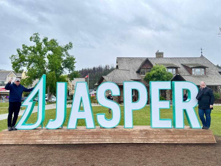 Two adults posing at the large turquoise JASPER sign in a small town park on a cloudy day.