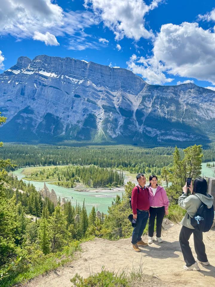 Couple posing for a photo at a cliffside viewpoint overlooking a winding turquoise river and vast pine forest.