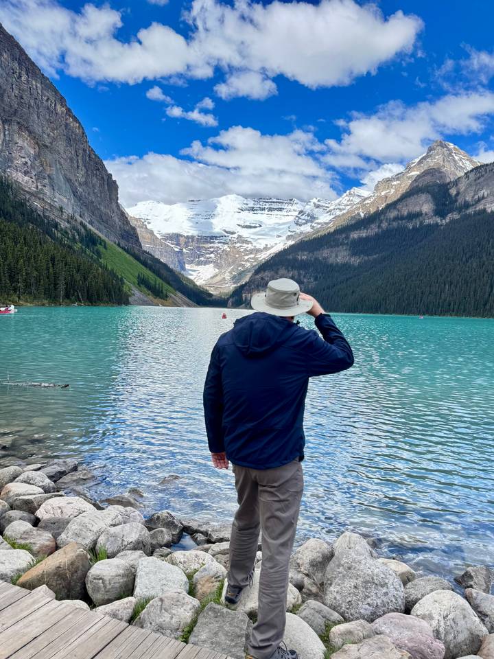 Lone traveller in sun hat gazing across vivid teal Lake Louise toward distant glacier and peaks.