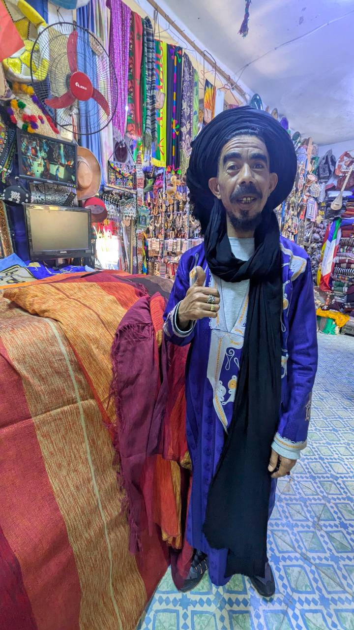 Young Berber man in a vibrant shop gives a thumbs-up, draped in indigo scarf and purple robe