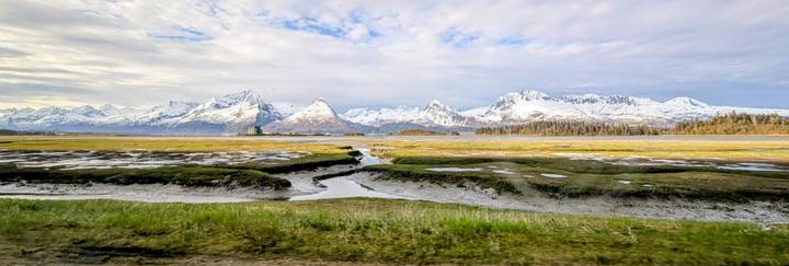 Snow-capped coastal mountains rise beyond tidal flats crossed by winding channels under a partly cloudy sky in Alaska.
