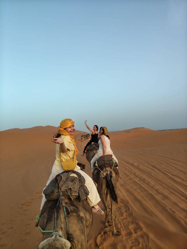 Smiling travellers riding camels across orange Sahara dunes, one waving at camera