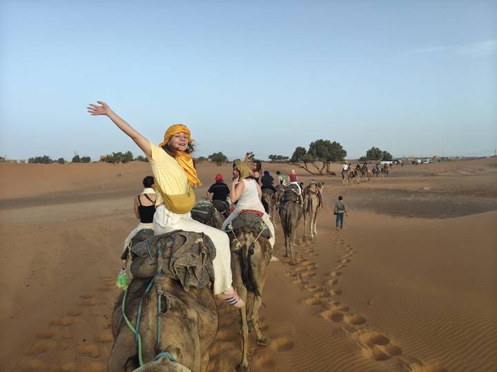 Long camel caravan of tourists winding through sandy landscape, lead rider with arms raised in joy