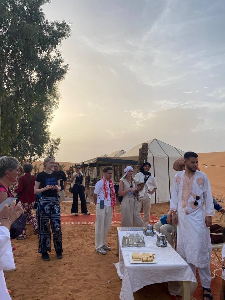 Travellers gather outside canvas tents at a Sahara desert camp in soft evening light