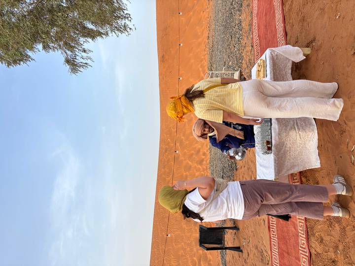 Berber host pours tea for two tourists at a low table set on desert sand