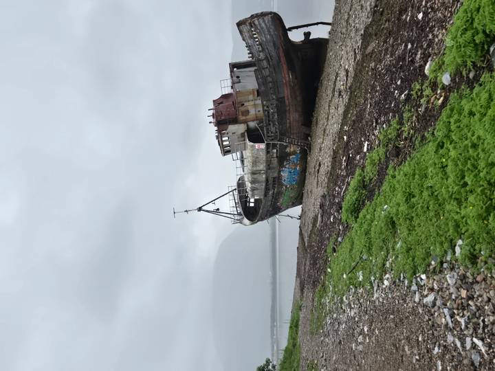 A rusted abandoned fishing boat rests on a pebbled shore under a grey, misty Highland sky.