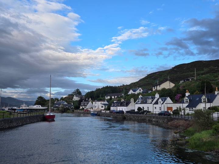 Charming white-washed cottages line a calm inlet with moored boats under a clearing sky.