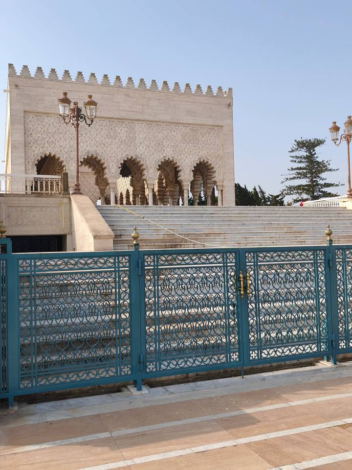 Staircase and arched colonnade of the Mausoleum of Mohammed V viewed behind decorative teal railings.