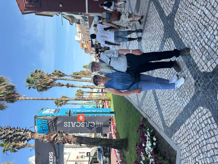 Couple stands on mosaic promenade in Cascais beside event banners and tall palm trees.
