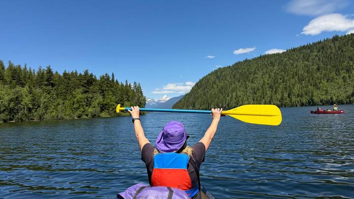 Kayaker raising a yellow paddle triumphantly while floating on a calm forested lake with snow-capped peaks in the distance.