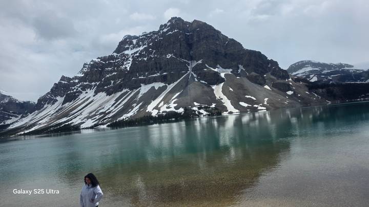 Snow-streaked mountain rising above a cold turquoise lake with lone person near shore; phone watermark text on lower left.