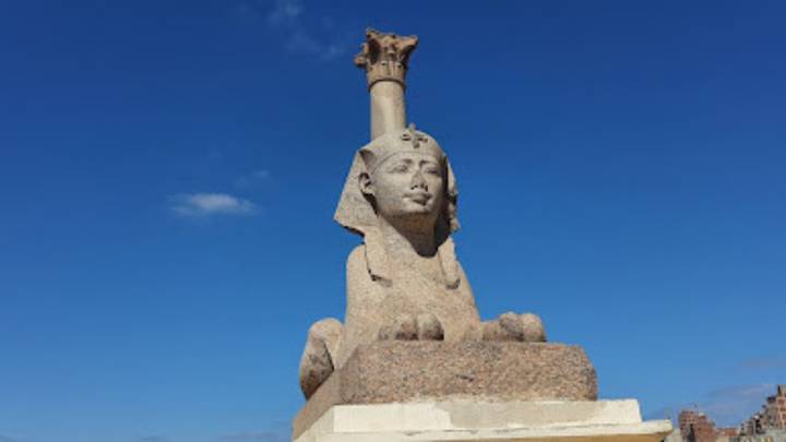 Granite sphinx statue atop a pedestal set against a cloudless blue sky.
