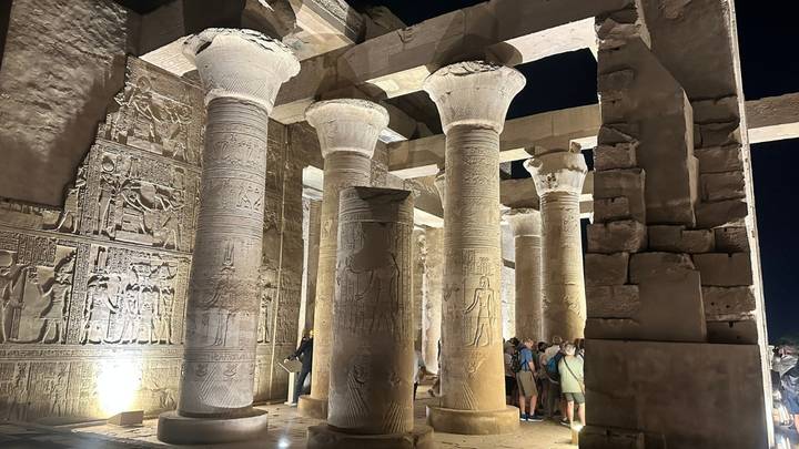 Massive stone columns with carved capitals inside Edfu temple lit at night while visitors look up.