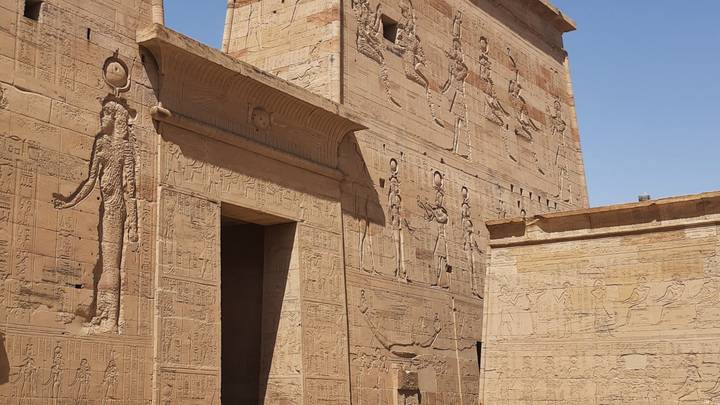 Sandstone walls of Philae Temple covered in large reliefs stand under bright blue sky.