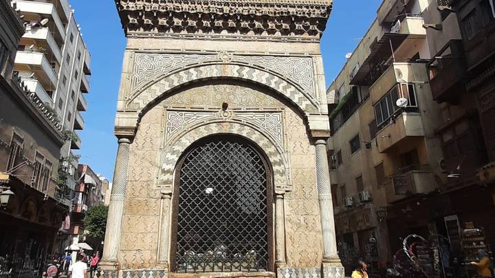 Ornate Mamluk sabil-kuttab with intricate stonework and iron grille set amid Cairo street buildings.