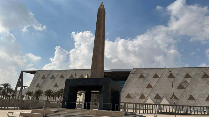 Modern museum facade with central obelisk framed by patterned stone walls and palm trees
