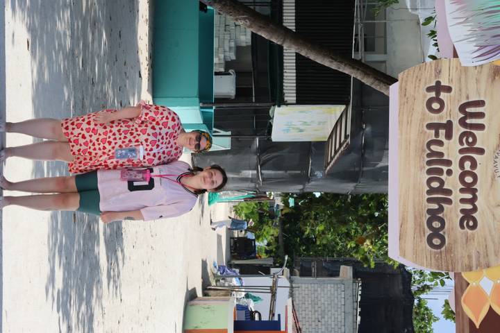 Two visitors pose beneath a large sign reading 'Welcome to Fulidhoo' on a sandy village street.