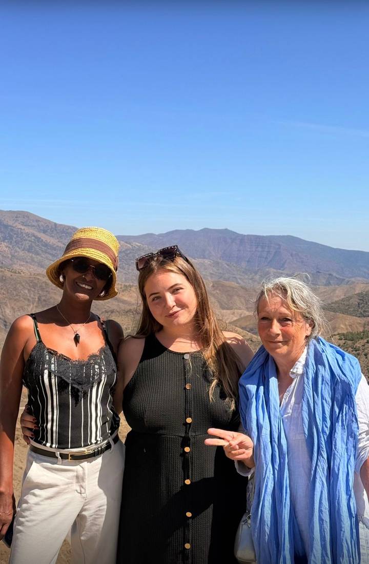 Three travelers smile in front of rugged mountain scenery under a bright blue sky.