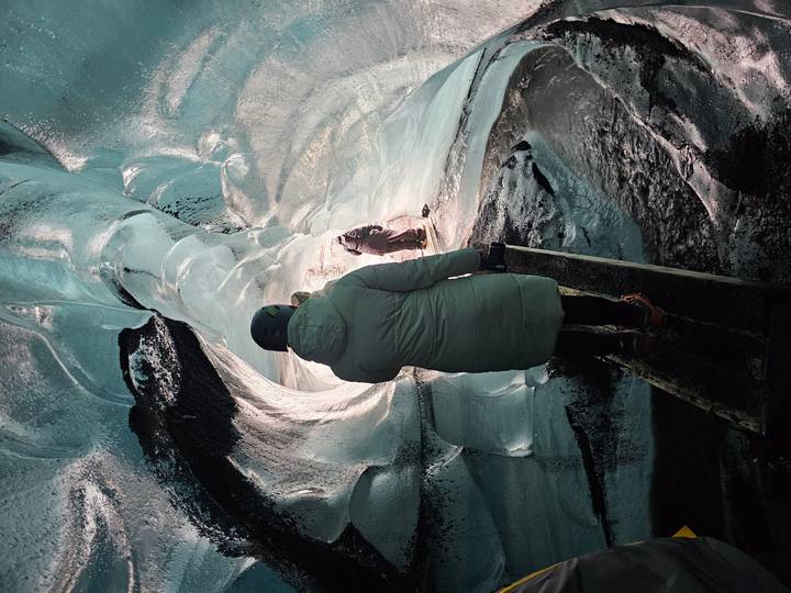 Traveler in parka and helmet walks a wooden plank through a turquoise ice tunnel