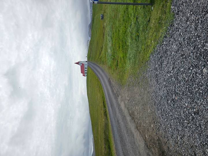 Isolated white church with red roof atop a grassy hill under a gray sky, reached by gravel road