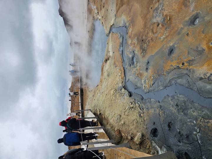 Visitors walk boardwalks around steaming, colorful geothermal pools and barren earth