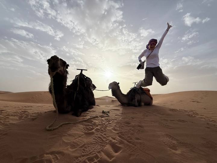 Joyful traveler jumps beside two resting camels on golden desert dunes at sunrise