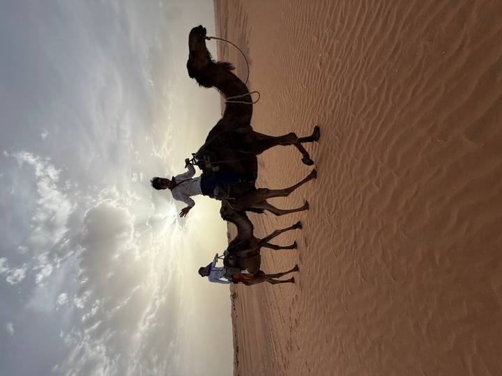 Two riders atop camels trek across rippled sand with hazy sun overhead