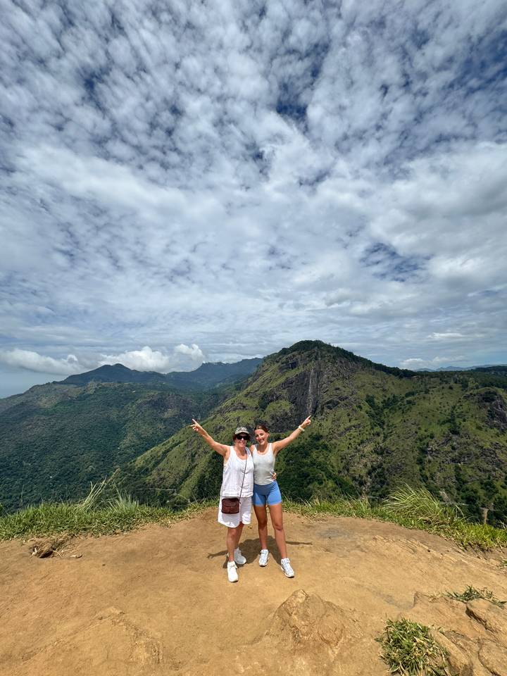 Two smiling women raising their arms on a mountain viewpoint with lush Ella hills behind