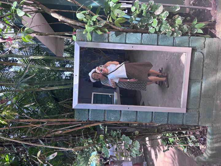 Mirror selfie of two women in a tropical garden with palm fronds and brick wall