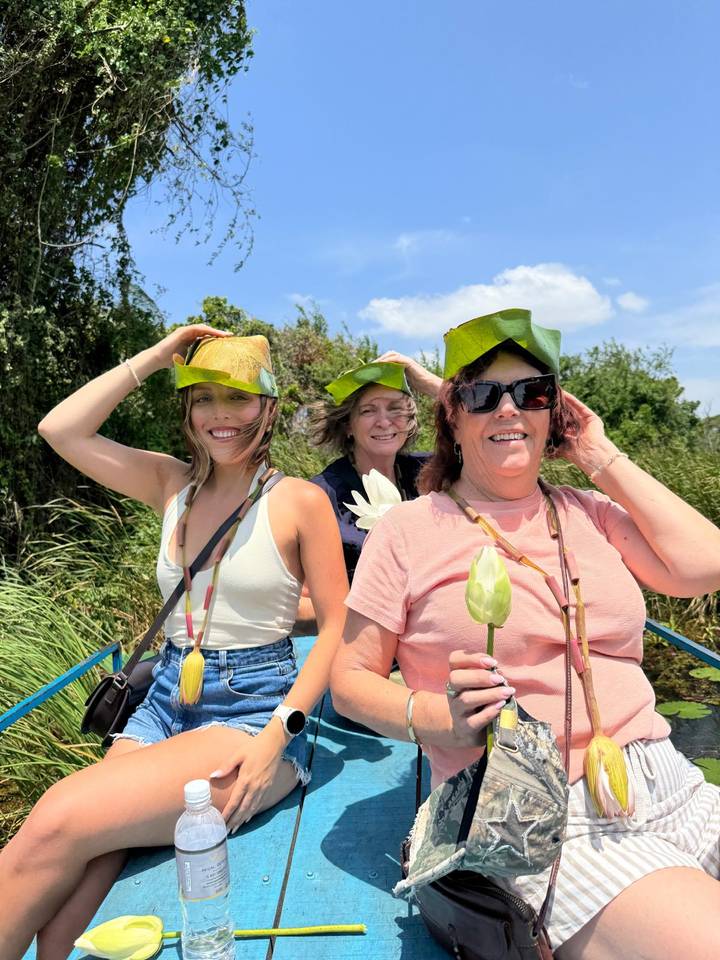 Three women smiling on a small boat wearing handmade leaf hats and holding lotus flowers