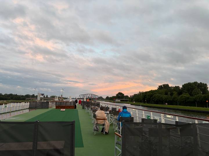 Passengers sit on deck chairs of a river cruise ship under a pastel evening sky with a distant bridge