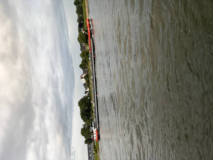 Long cargo barge travels along a wide river under a cloudy sky with shoreline houses behind