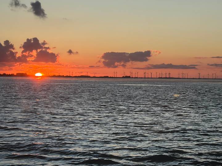 Vibrant orange sunset over open water with silhouettes of distant wind turbines and low clouds