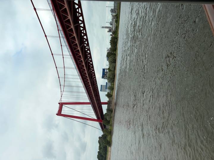 Red steel suspension bridge spans a broad river on a cloudy day