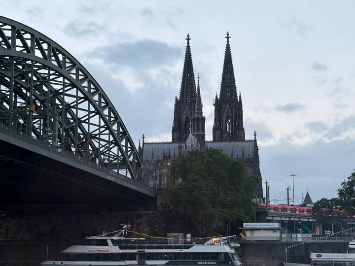 View of Cologne Cathedral’s twin spires beside the steel arches of Hohenzollern Bridge under a grey evening sky.