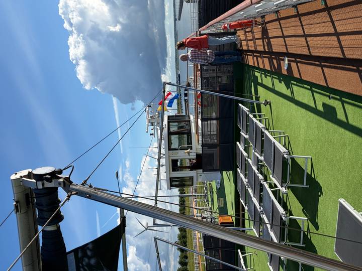 Sunlit upper deck of a river cruise ship with lounge chairs, flag mast and two passengers enjoying the view of open water.