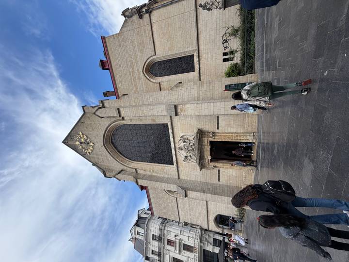 Stone church with a large arched window and clock above the entrance as visitors file through the doorway on a bright day.
