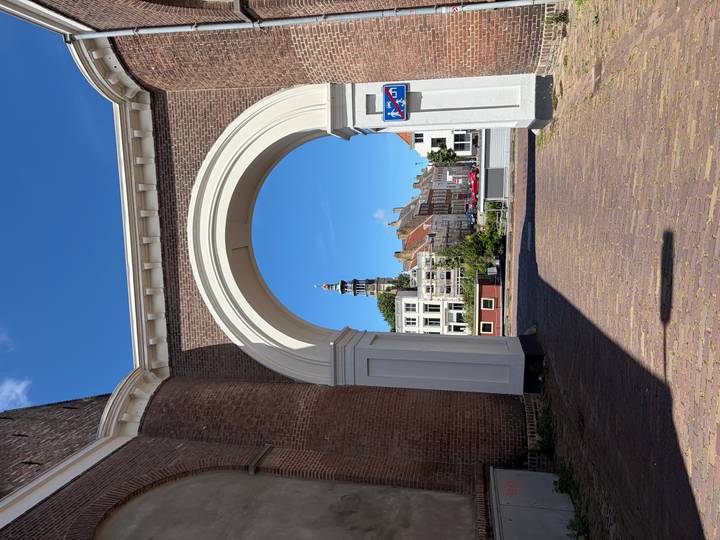White stone archway framing a distant Dutch clock tower and traditional brick houses under clear blue sky.