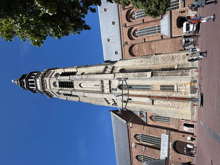 Tall medieval church bell tower rises against a vivid blue sky as two pedestrians walk across the square below.