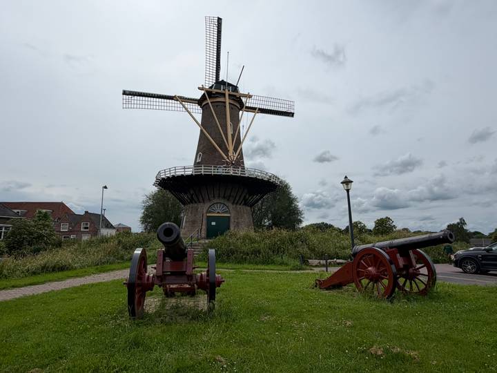 Historic Dutch windmill surrounded by grassy field and two old cannons pointing outward under a grey sky.