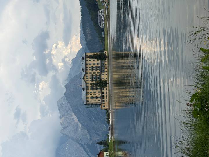 Grand yellow lakeside hotel reflected in calm waters with towering mountains behind under a moody sky.